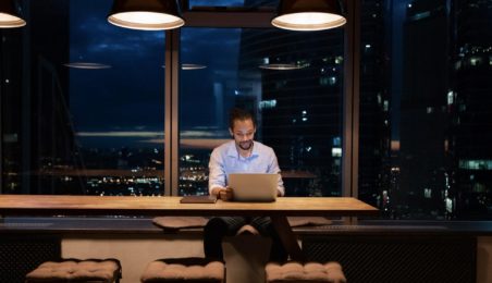 Venture capital firm employee (man) working on laptop, dark sky behind him out the window